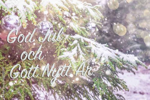 Text in Swedish "God jul och gott nytt ar"means "Merry Christmas and Happy New Year". Greetings in Swedish. Blurred background of fir tree in snow decorated with silver lights, toys. Bokeh. Postcard. 