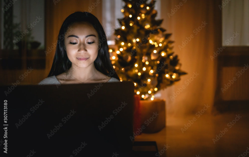 Happy asian girl shopping online with laptop sitting near decorated christmas tree background at evening Happy asian girl shopping online with laptop sitting near decorated christmas tree background at evening