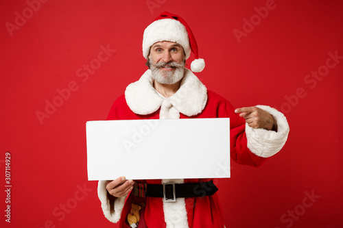 Elderly gray-haired mustache bearded Santa man in Christmas hat posing isolated on red background. New Year 2020 celebration holiday concept. Mock up copy space. Pointing on white blank sign board.