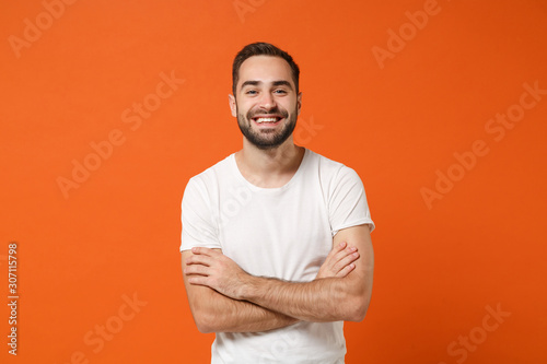 Smiling funny young man in casual white t-shirt posing isolated on bright orange wall background, studio portrait. People sincere emotions lifestyle concept. Mock up copy space. Holding hands crossed.
