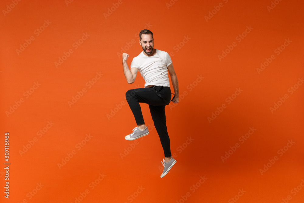 Happy joyful young man in casual white t-shirt posing isolated on bright orange wall background studio portrait. People lifestyle concept. Mock up copy space. Having fun jumping, doing winner gesture. Happy joyful young man in casual white t-shirt posing isolated on bright orange wall background studio portrait. People lifestyle concept. Mock up copy space. Having fun jumping, doing winner gesture.