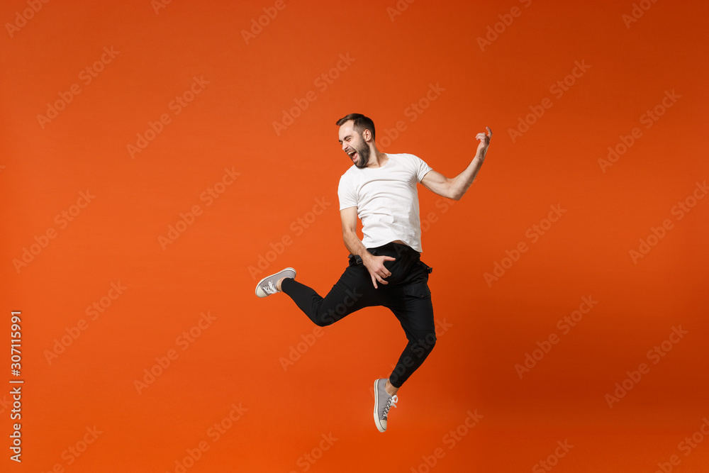 Crazy young man in casual white t-shirt posing isolated on orange wall background studio portrait. People lifestyle concept. Mock up copy space. Having fun, jumping fooling around like playing guitar.