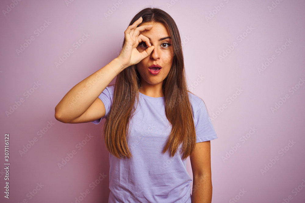 Young beautiful woman wearing casual t-shirt standing over isolated pink background doing ok gesture shocked with surprised face, eye looking through fingers. Unbelieving expression.