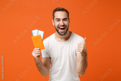 Funny young man in casual white t-shirt posing isolated on orange background studio portrait. People lifestyle concept. Mock up copy space. Holding passport boarding pass ticket, showing thumb up.