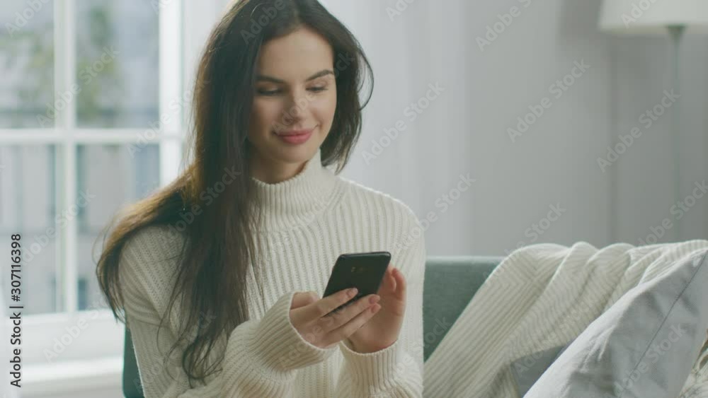 Young Woman Using Smartphone, Drinks Tea while Sitting on the Chair. Sensual Girl Wearing Sweater Surfs Internet, Posts On Social Media, Shares Pictures while Relaxing in Cozy Apartment Young Woman Using Smartphone, Drinks Tea while Sitting on the Chair. Sensual Girl Wearing Sweater Surfs Internet, Posts On Social Media, Shares Pictures while Relaxing in Cozy Apartment