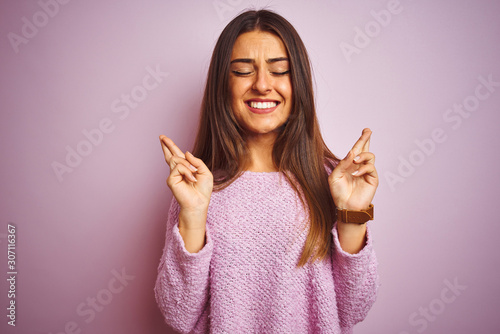Young beautiful woman wearing casual sweater standing over isolated pink background gesturing finger crossed smiling with hope and eyes closed. Luck and superstitious concept.