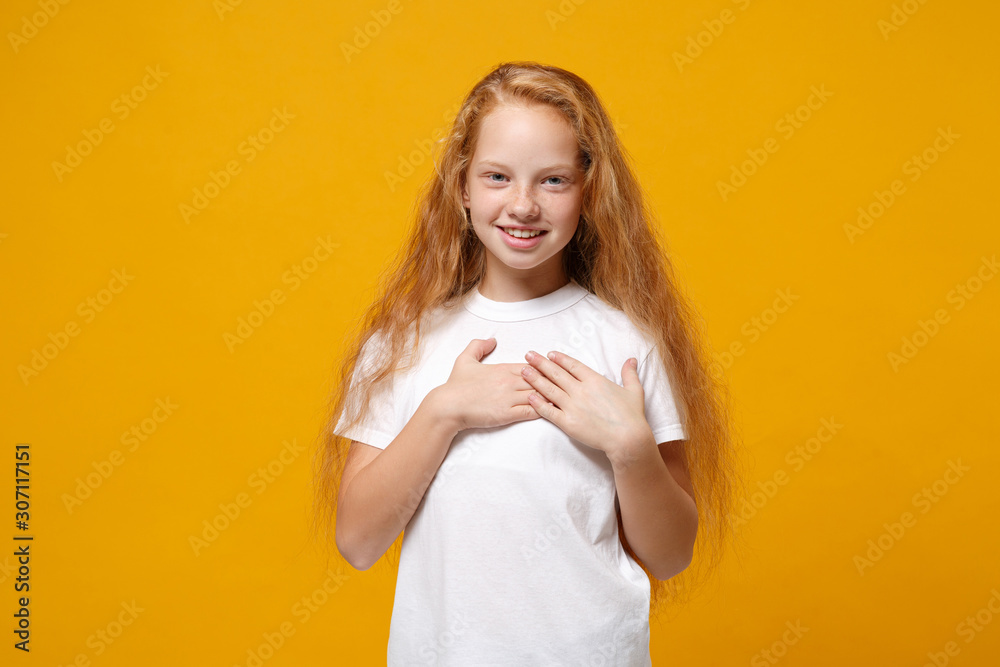 Smiling little ginger kid girl 12-13 years old in white t-shirt isolated on yellow wall background children studio portrait. Childhood lifestyle concept. Mock up copy space. Holding hands on heart. Smiling little ginger kid girl 12-13 years old in white t-shirt isolated on yellow wall background children studio portrait. Childhood lifestyle concept. Mock up copy space. Holding hands on heart.