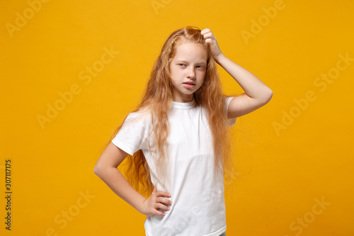 Pensive little ginger kid girl 12-13 years old in white t-shirt isolated on yellow background children portrait. Childhood lifestyle concept. Mock up copy space. Putting hand on head, looking aside.
