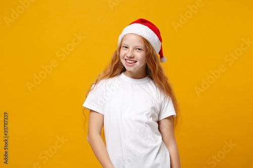 Smiling little ginger kid Santa girl 12-13 years old in white t-shirt, Christmas hat isolated on yellow background. Happy New Year 2020 celebration holiday concept. Mock up copy space. Looking camera. © ViDi Studio