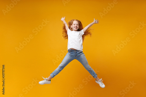 Funny little ginger kid girl 12-13 years old in white t-shirt isolated on yellow background. Childhood lifestyle concept. Mock up copy space. Having fun, fooling around, jumping, spreading hands legs.