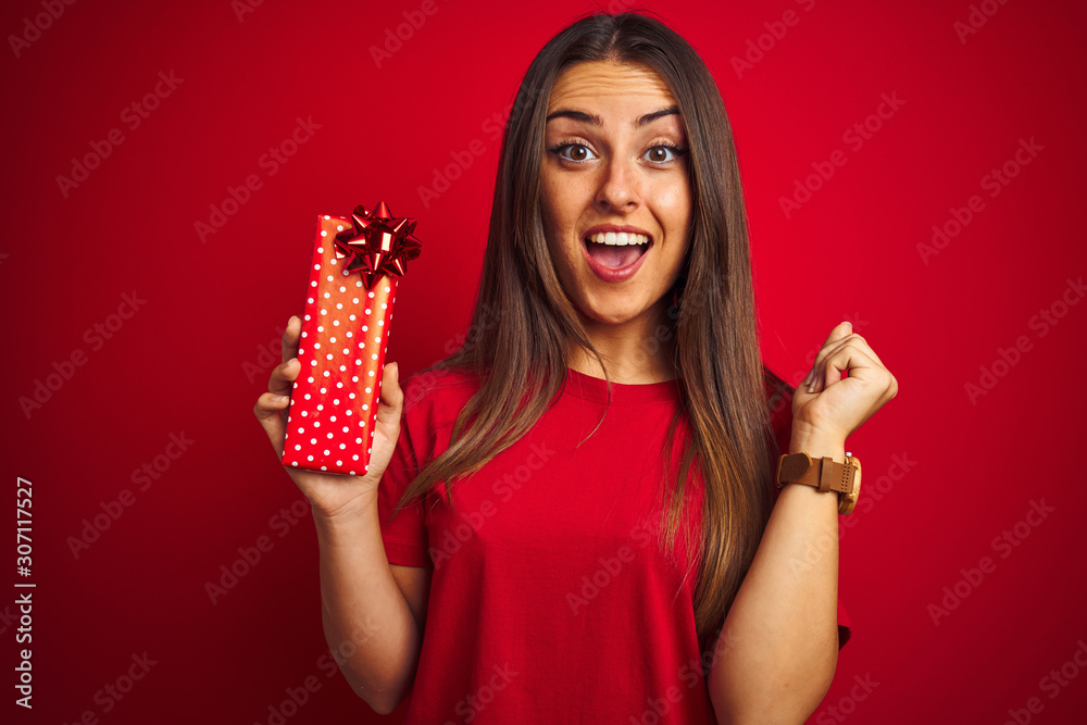 Young beautiful woman holding birthday gift standing over isolated red background screaming proud and celebrating victory and success very excited, cheering emotion Young beautiful woman holding birthday gift standing over isolated red background screaming proud and celebrating victory and success very excited, cheering emotion