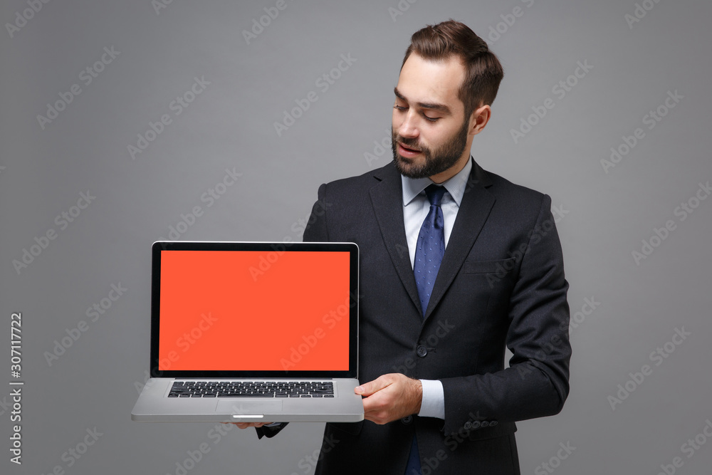 Young business man in classic black suit shirt tie posing isolated on grey background. Achievement career wealth business concept. Mock up copy space. Hold laptop pc computer with blank empty screen. Young business man in classic black suit shirt tie posing isolated on grey background. Achievement career wealth business concept. Mock up copy space. Hold laptop pc computer with blank empty screen.