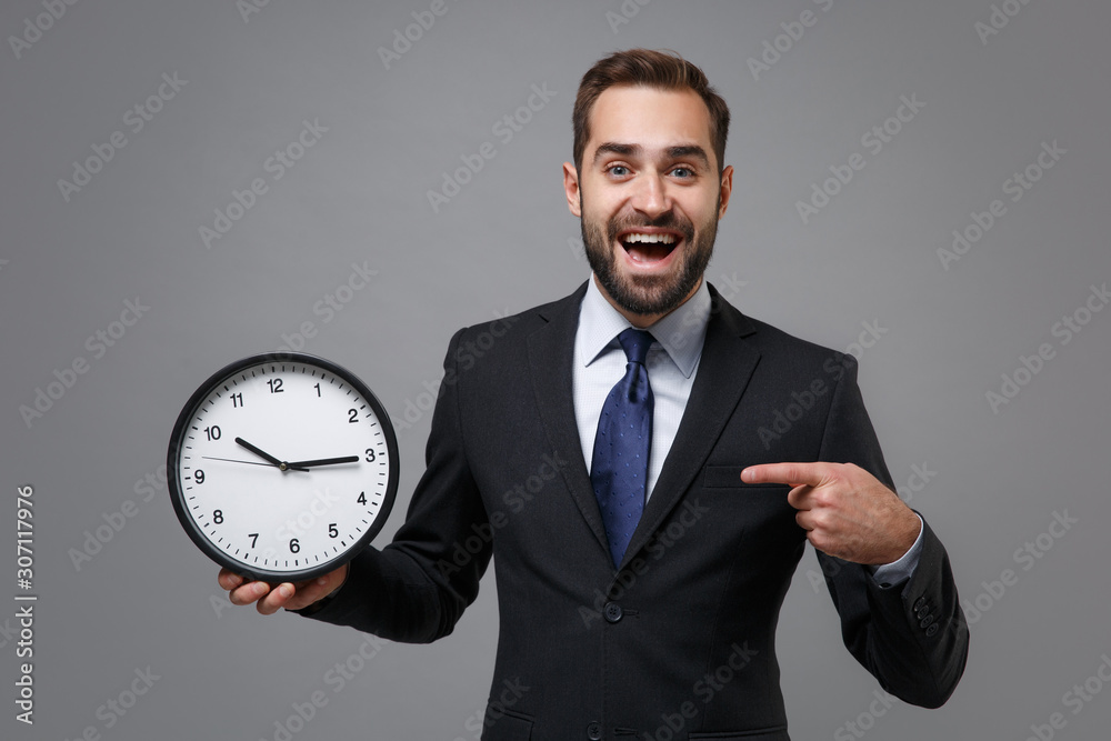 Excited young bearded business man in classic black suit shirt tie posing isolated on grey background. Achievement career wealth business concept. Mock up copy space. Pointing index finger on clock. Excited young bearded business man in classic black suit shirt tie posing isolated on grey background. Achievement career wealth business concept. Mock up copy space. Pointing index finger on clock.