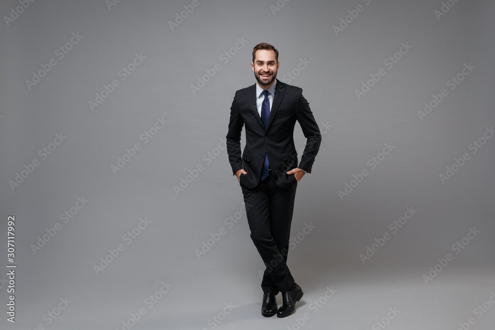 Smiling young bearded business man in classic black suit shirt tie posing isolated on grey wall background. Achievement career wealth business concept. Mock up copy space. Holding hands in pockets. Smiling young bearded business man in classic black suit shirt tie posing isolated on grey wall background. Achievement career wealth business concept. Mock up copy space. Holding hands in pockets.