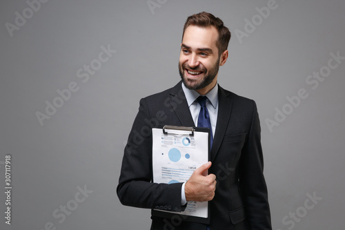 Cheerful young business man in suit shirt tie posing isolated on grey background. Achievement career wealth business concept. Mock up copy space. Hold clipboard with papers document, looking aside.