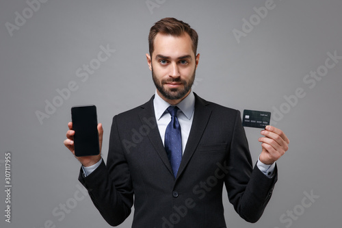 Young business man in suit shirt tie posing isolated on grey background. Achievement career wealth business concept. Mock up copy space. Holding mobile phone with blank empty screen, credit bank card.