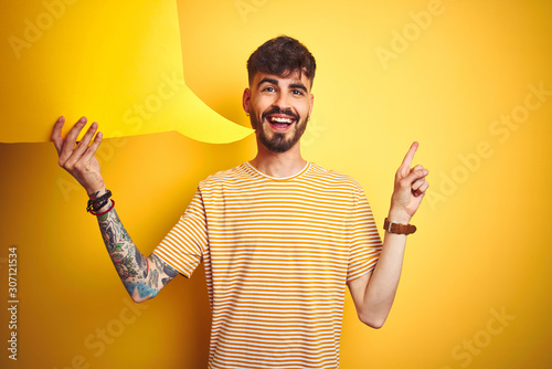 Young man with tattoo holding speech bubble standing over isolated yellow background very happy pointing with hand and finger to the side