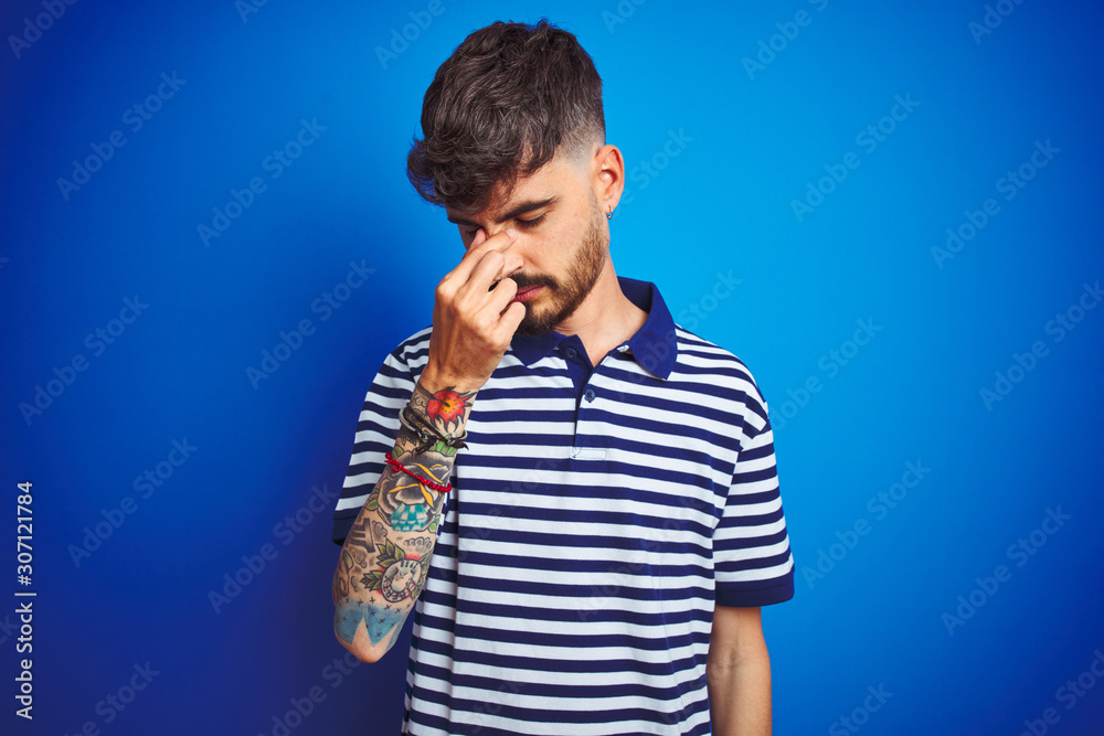 Young man with tattoo wearing striped polo standing over isolated blue background tired rubbing nose and eyes feeling fatigue and headache. Stress and frustration concept. Young man with tattoo wearing striped polo standing over isolated blue background tired rubbing nose and eyes feeling fatigue and headache. Stress and frustration concept.