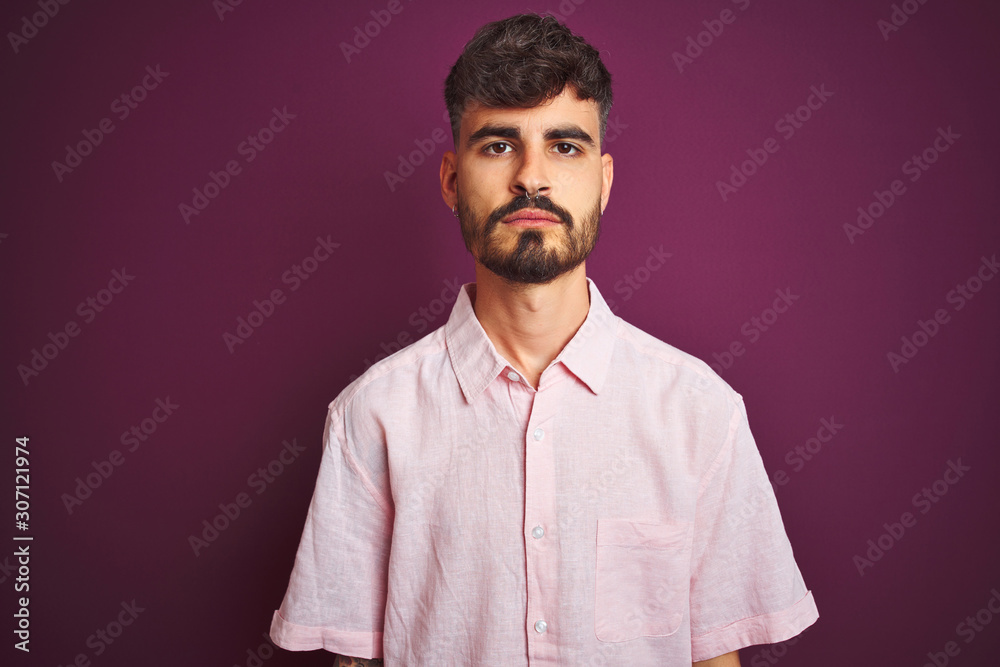Young man with tattoo wearing summer pink shirt standing over isolated purple background with serious expression on face. Simple and natural looking at the camera. Young man with tattoo wearing summer pink shirt standing over isolated purple background with serious expression on face. Simple and natural looking at the camera.