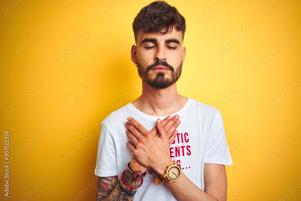 Young man with tattoo wearing fanny t-shirt standing over isolated yellow background smiling with hands on chest with closed eyes and grateful gesture on face. Health concept. Young man with tattoo wearing fanny t-shirt standing over isolated yellow background smiling with hands on chest with closed eyes and grateful gesture on face. Health concept.