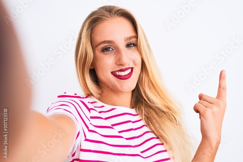 Beautiful woman wearing striped t-shirt make selfie by camera over isolated white background very happy pointing with hand and finger to the side