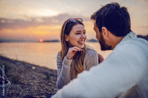 Cute smiling Caucasian brunette sitting on coast near river with her loving boyfriend and flirting. In background is sunset. photo