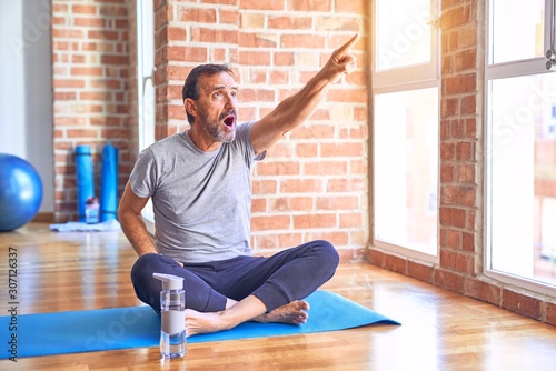 Fototapeta Naklejka Na Ścianę i Meble -  Middle age handsome sportman sitting on mat doing stretching yoga exercise at gym Pointing with finger surprised ahead, open mouth amazed expression, something on the front