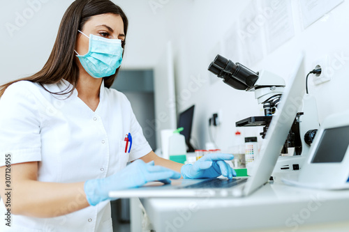 Tableau sur toile Caucasian lab assistant with protective mask and rubber gloves sitting in lab and typing test results on laptop