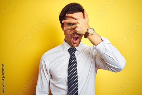 Young handsome businessman wearing elegant shirt and tie over isolated yellow background peeking in shock covering face and eyes with hand, looking through fingers with embarrassed expression.