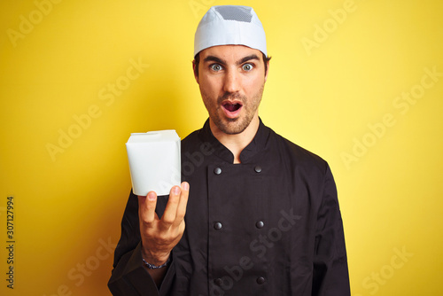 Young chef man wearing uniform and hat holding delivery box over isolated yellow background scared in shock with a surprise face, afraid and excited with fear expression