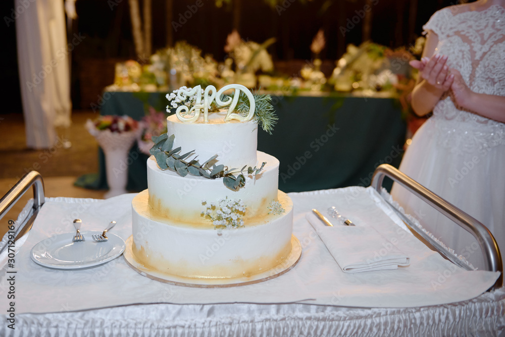 Elegant white wedding cake decorated with flowers, green leaves and golden glitter on table in restaurant, copy space
