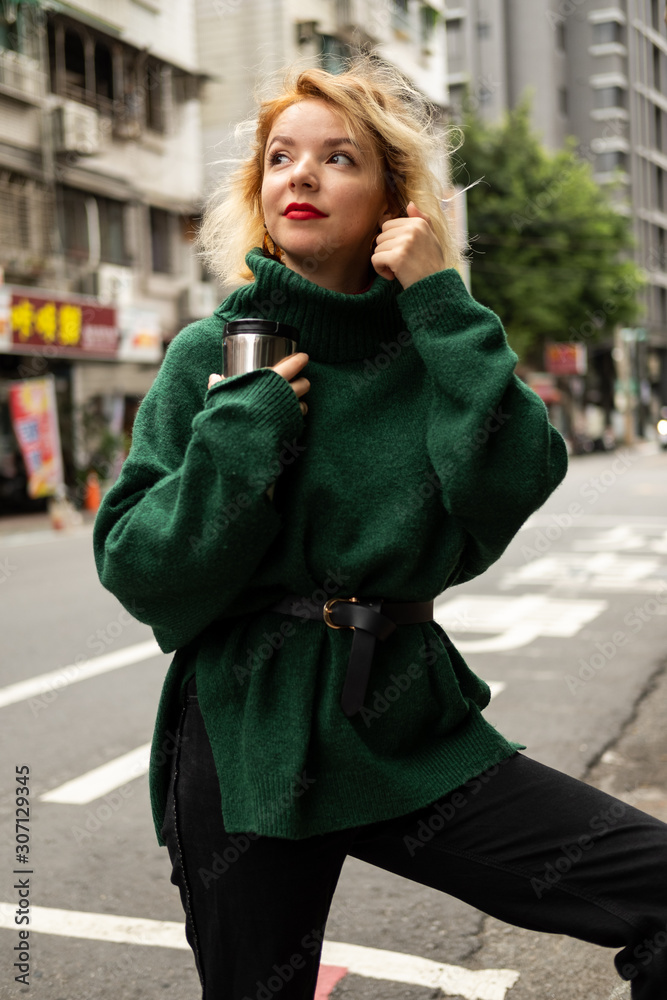Fototapeta premium pretty young blond smiling girl in green sweater on the street hold cup in her hands outside in day time