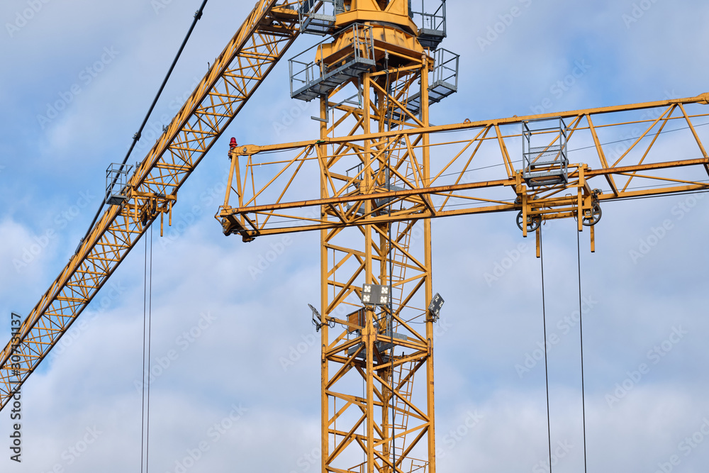 Two yellow big cranes with their arms against a bright sky with clouds on a big construction site.