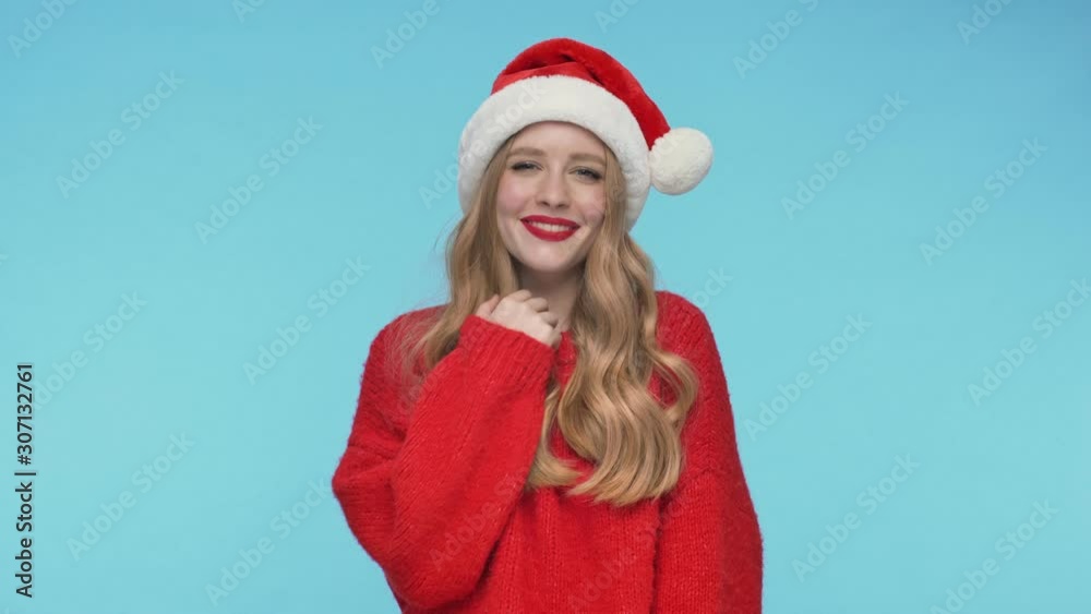 Cheerful shy pretty woman in christmas hat becoming touched and covering her face with arm and hair over turquoise background Cheerful shy pretty woman in christmas hat becoming touched and covering her face with arm and hair over turquoise background