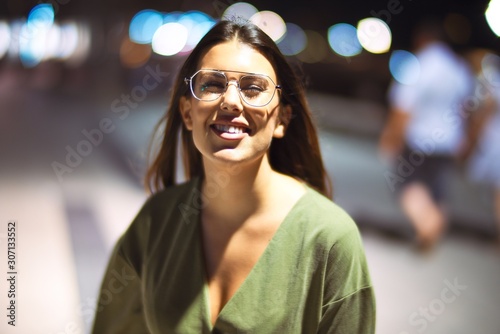 Young beautiful girl smiling happy and confident walking at the promenade, standing with a smile on face around night lights bokeh
