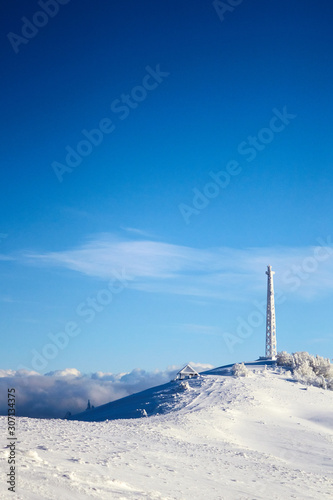 Icy cellular base station antenna covered with snow. Cell site tower on moutain hill. Telephone network transceivers and communication equipment on frosty weather day after snowstorm. Blue sky.