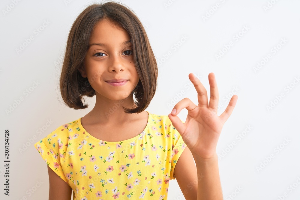 Young beautiful child girl wearing yellow floral dress standing over isolated white background doing ok sign with fingers, excellent symbol Young beautiful child girl wearing yellow floral dress standing over isolated white background doing ok sign with fingers, excellent symbol