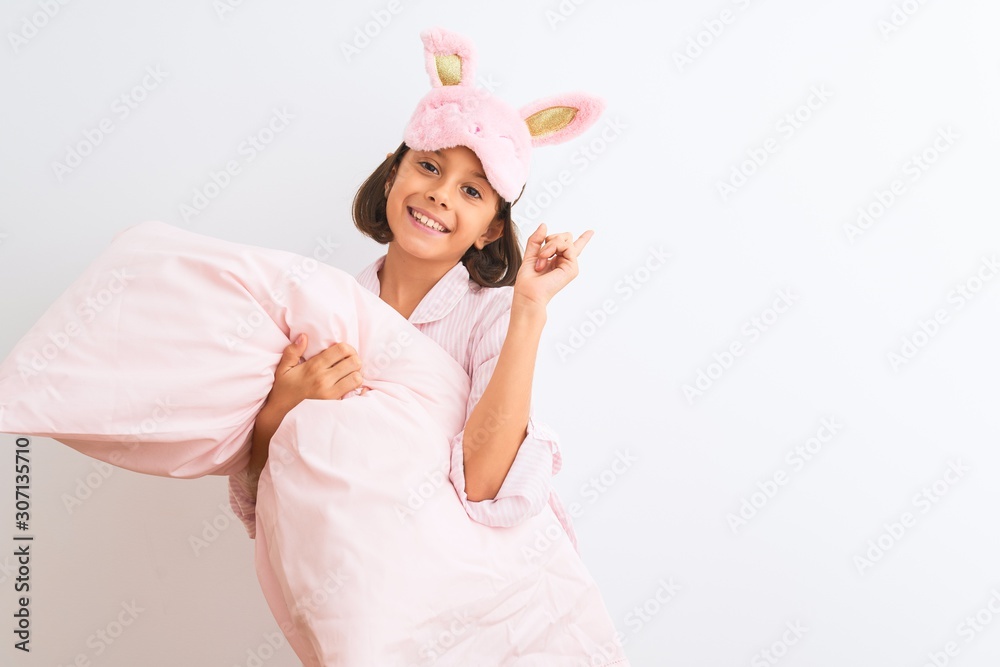 Child girl wearing sleep mask and pajama holding pillow over isolated white background surprised with an idea or question pointing finger with happy face, number one