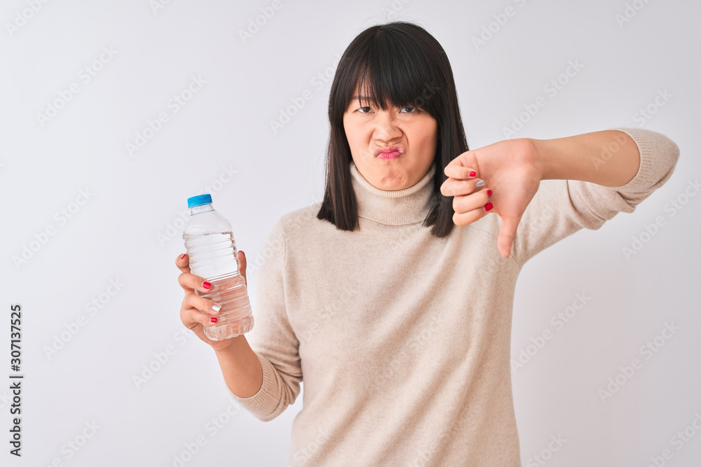 Young beautiful Chinese woman holding bottle of water over isolated white background with angry face, negative sign showing dislike with thumbs down, rejection concept Young beautiful Chinese woman holding bottle of water over isolated white background with angry face, negative sign showing dislike with thumbs down, rejection concept