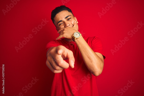 Young brazilian man wearing t-shirt standing over isolated red background laughing at you, pointing finger to the camera with hand over mouth, shame expression