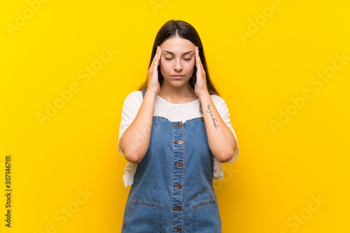 Young woman in dungarees over isolated yellow background with headache