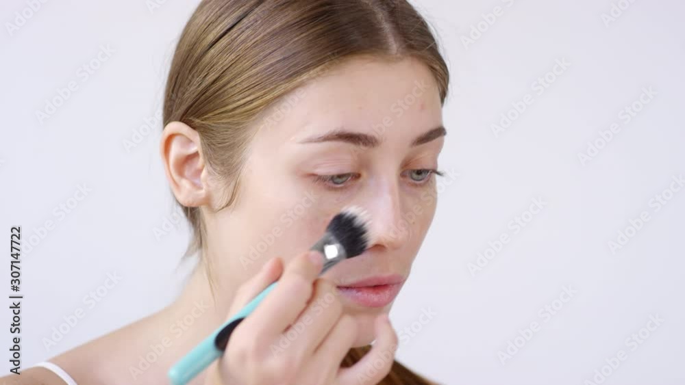 Close up shot with grey background. Young beautiful woman using brush and applying light primer to her face while doing her makeup Close up shot with grey background. Young beautiful woman using brush and applying light primer to her face while doing her makeup
