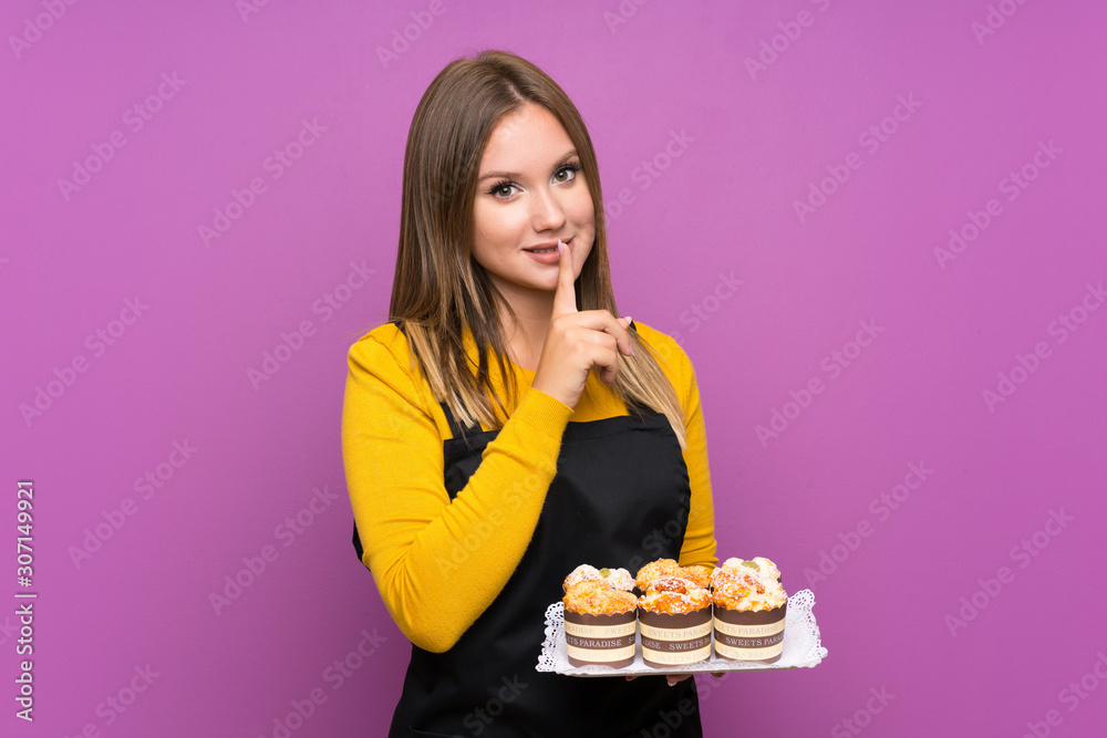 Teenager girl holding lots of different mini cakes over isolated purple background doing silence gesture Teenager girl holding lots of different mini cakes over isolated purple background doing silence gesture