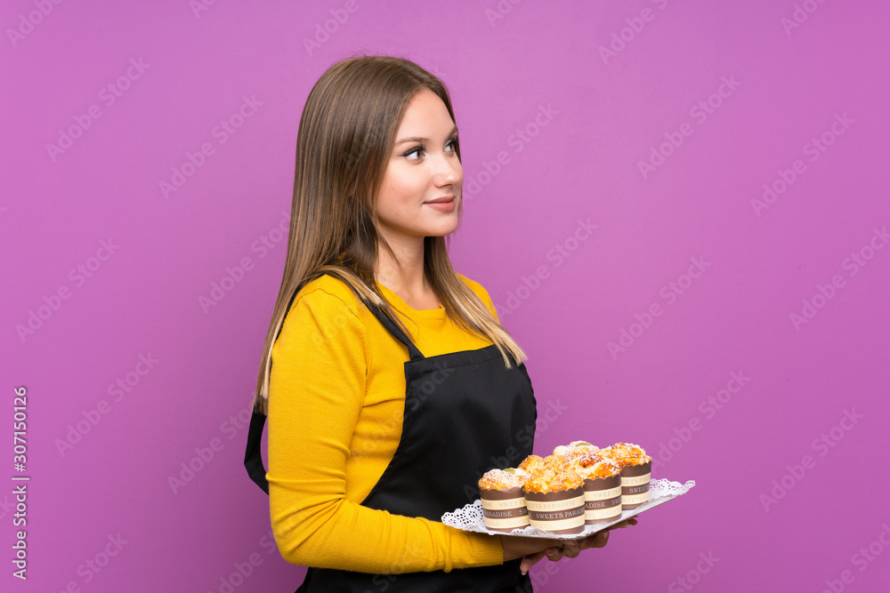Teenager girl holding lots of different mini cakes over isolated purple background looking to the side Teenager girl holding lots of different mini cakes over isolated purple background looking to the side