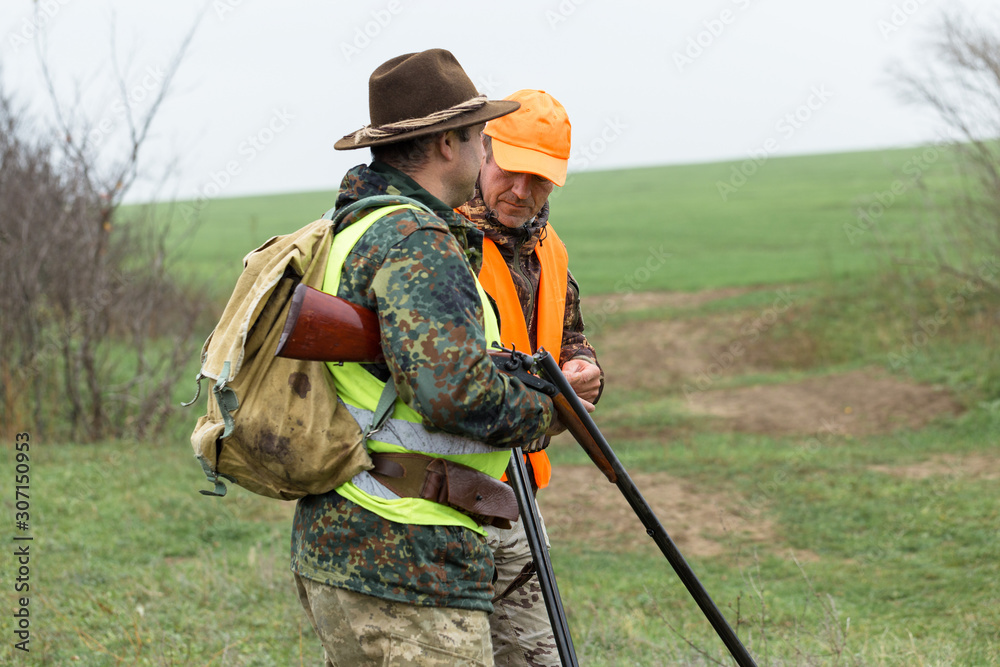 Fototapeta premium A mans with a gun in his hands and an orange vest on a pheasant hunt in a wooded area in cloudy weather. Hunters with dogs in search of game.