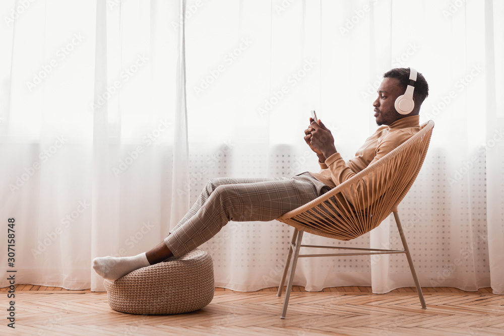 Millennial Afro Guy In Headphones Using Smartphone Sitting Indoor - obrazy, fototapety, plakaty 