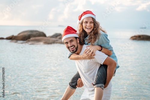 Young beautiful couple in Santa Claus hats walks along the shore of the tropical sea at the resort, vacation and travel for the New Year and Christmas time