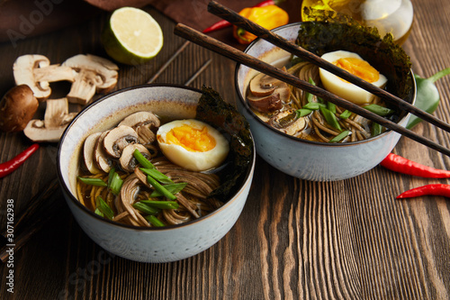 traditional spicy ramen in bowls with chopsticks and vegetables on wooden table with napkin