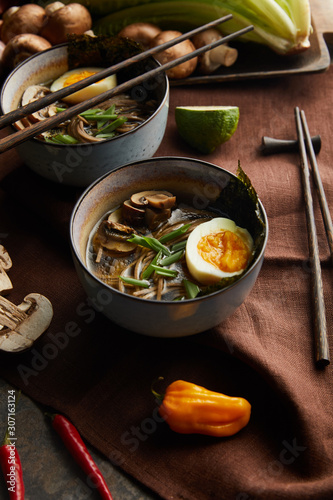 traditional spicy ramen in bowls with chopsticks and vegetables on brown napkin on stone surface