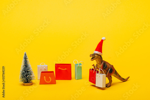 Fototapeta Naklejka Na Ścianę i Meble -  Toy dinosaur in santa hat with shopping bags and christmas tree on yellow background